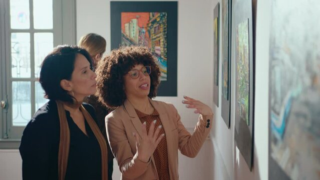 Two female friends walking around art gallery, observing paintings collection and having discussion while visiting fine art exhibition