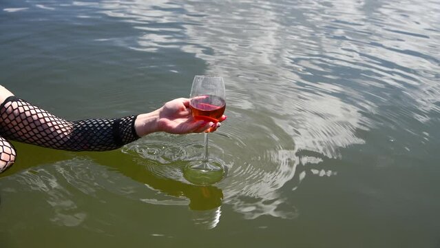 Sexy plus size caucasian woman wearing mesh crop top and standing in water raises wineglass with red wine over water. Mature content. Real time video. Adult leisure theme.