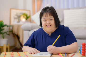 Asian fat woman with Down syndrome smiling looking at camera Sitting at the drawing table and taking notes in a workbook with colored pencils in the living room at home