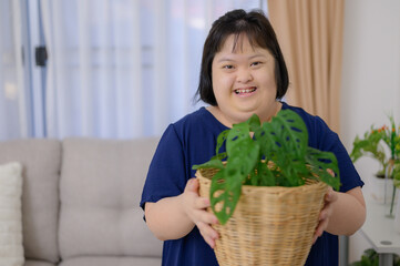 A young Asian woman with Down syndrome smiling and looking at the camera. Holding a potted plant in the living room at home
