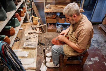 High angle of mature experienced shoemaking master in casualwear sitting in front of table and...