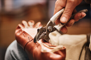 Metallic pliers in hand of male shoemaker fixing upper part of boot to sole while sitting in front...