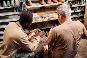 Young African American apprentice of shoemaker fixing upper part of boot to sole while sitting next...