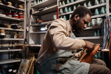 Young African American male owner of shoemaking workshop fixing upper part of unfinished boot to...