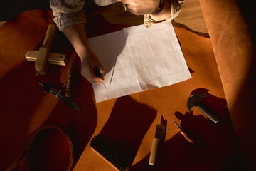 Craftsman drawing scheme of future wallet, creating leatherwork at studio. Male tanner at working...