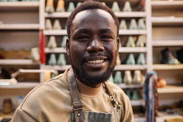 Happy young African American shoemaker or apprentice looking at camera while standing in spacious workshop with supplies