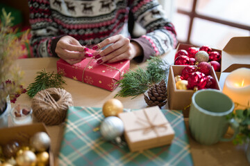 Young asian woman in sweater wrapping christmas gift and decorating with hemp rope while preparing toys ornaments and presents to celebrate for christmas festive holiday and winter seasons at house