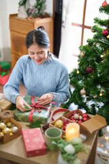 Young asian woman in sweater wrapping christmas gift to tie red ribbon decorating with pine branch and berries while preparing presents to celebrate for christmas festive holiday and winter seasons