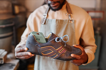 Cropped shot of young African American shoemaker in apron holding unfinished grey suede cross shoe...