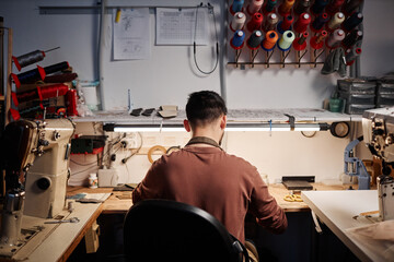 Rear view of young shoemaker in t-shirt sitting by table with supplies and handtools surrounded by...