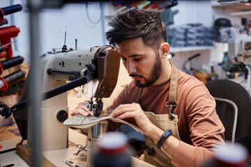 Young Asian man in workwear sitting in front of sewing machine in workshop and putting together...