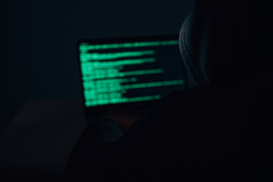 Behind the scenes of an anonymous hacker wearing a hoodie over his head in the dark hacking into a victim's financial system. Data thief, internet fraud, darknet and cyber security concept.
