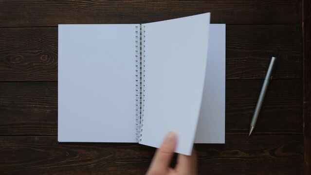 A woman's hand flips through the pages of a blank notepad on a spiral on a brown wooden table, view from the background