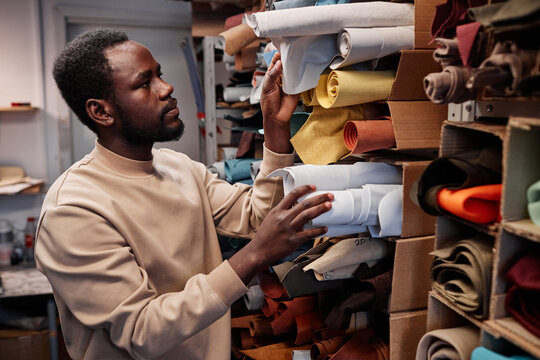 Young African American Male Leatherworker Choosing Suitable Color Of Suede Or Leather Textile For Creating New Items For Sale In Craft Shop