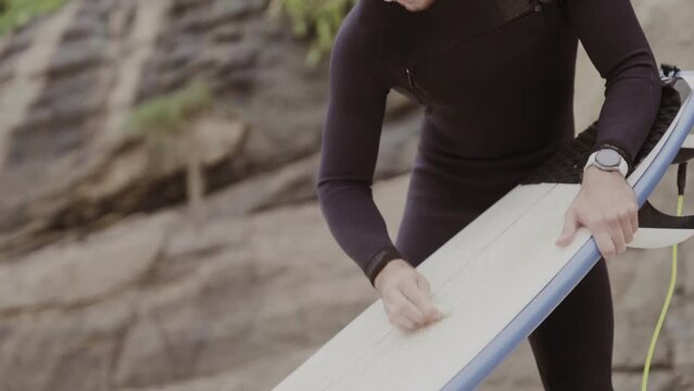Jovem na praia, concentrado, aplicando parafina na prancha de surf. Cinematico 4k.