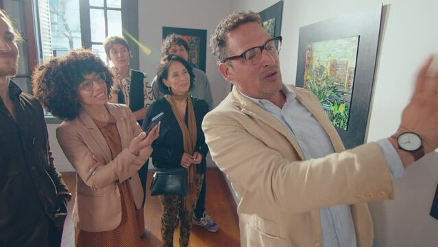 Charismatic mid-aged gallery guide giving giving speech about artwork presented on exhibition, attentive visitors listening to him during group tour