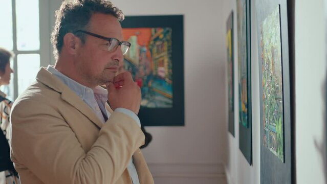 Mid-aged man scrutinizing painting and rubbing his chin thoughtfully in exhibition hall of art gallery