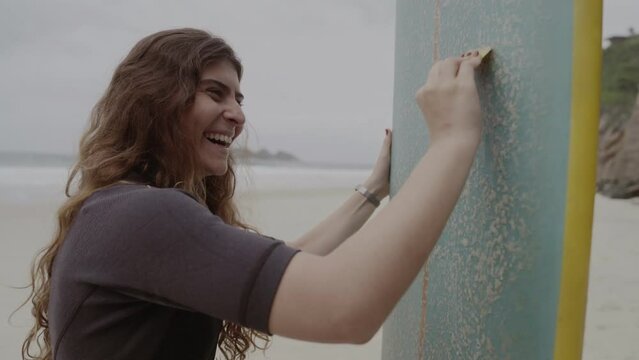Mulher jovem sorridente na praia aplicando parafina em sua prancha de surf. Cinematico 4k.