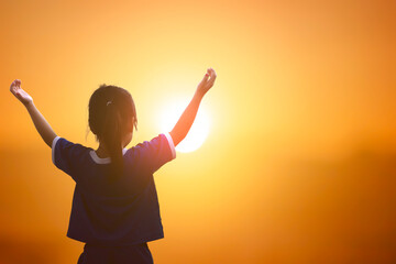 Silhouette of little girl raising hands on sunset sky background.
