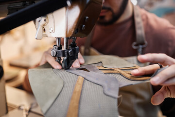 Close-up of young shoemaker or tanner using sewing machine while processing piece of grey suede and...