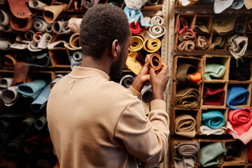 Back view of young African American male leatherworker standing by shelves with rolled suede and leather textile in workshop