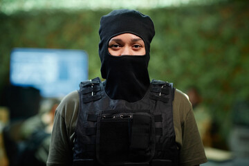Young serious female military officer in black balaclava hiding half of her face and protective body armor standing in front of camera