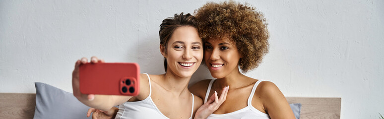 happy woman taking selfie on smartphone near african american girlfriend in bedroom, banner