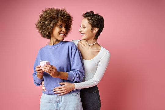 cheerful lesbian woman looking at happy african american girlfriend with present on Valentins day