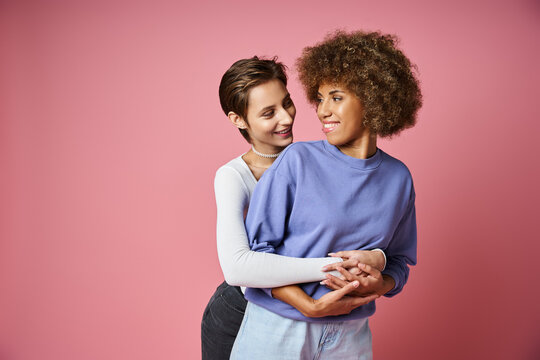 happy woman embracing her african american girlfriend on pink background, lgbtq couple love