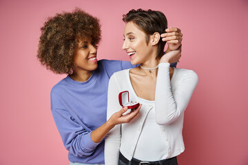 african american woman holding box with engagement ring near amazed girlfriend on pink background