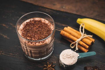 Milkshake smoothie, protein drink with grated chocolate in a glass on dark wooden board with bananas, protein powder in measuring spoon, star anise, almonds, cinnamon stick, chocolate pieces