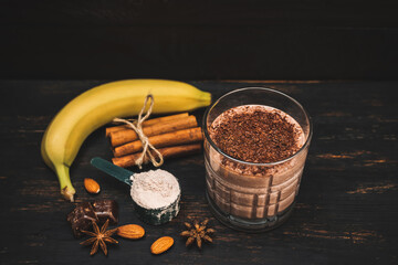 Milkshake smoothie, protein drink with grated chocolate in a glass on dark wooden board with bananas, protein powder in measuring spoon, star anise, almonds, cinnamon stick, chocolate pieces