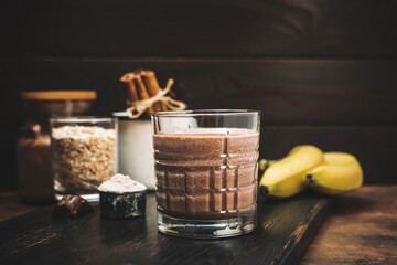 Chocolate milkshake smoothie, protein drink in a glass on dark wooden board with bananas, protein...