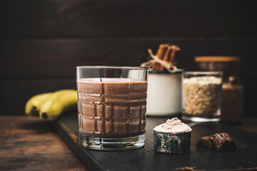 Chocolate milkshake smoothie, protein drink in a glass on dark wooden board with bananas, protein powder in measuring spoon and chocolate pieces
