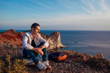 Tourist hiker having rest on mountain top by sea on Santorini island enjoying landscape. Happy man admires sunset