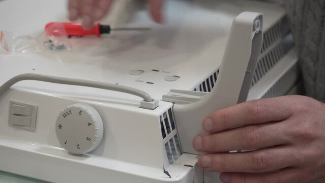 Man mounting a plastic legs on an electric heate, a man with a screwdriver installs plastic legs on a household electric convector