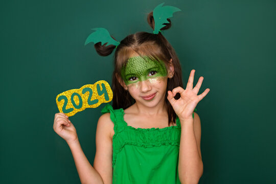 A child dressed up as a dragon with gold sequins on his face holds the numbers of 2024 and makes a gesture with his fingers ok