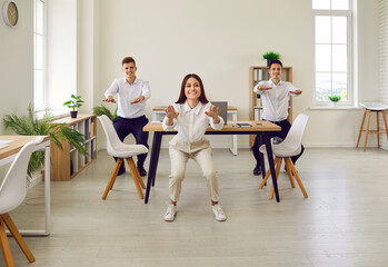 Full length portrait of young business people doing stretching sit-ups exercises standing at workplace in office. Group of smiling company employees exercising for health during a break from a work. - obrazy, fototapety, plakaty