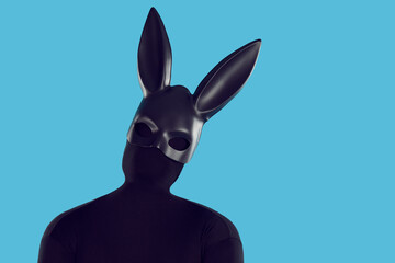 Studio portrait fashion model disguised in rabbit costume. Anonymous adult man in faceless monochromatic black bodysuit and long eared bunny mask tilts head isolated like silhouette on blue background