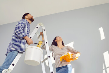 Happy young couple painting the ceiling of their new home holding paint rollers and standing on the ladder. Married man and woman doing repair renovation preparing to move into a new flat. - obrazy, fototapety, plakaty