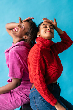 Two women in colorful attire striking playful and confident poses against a blue backdrop
