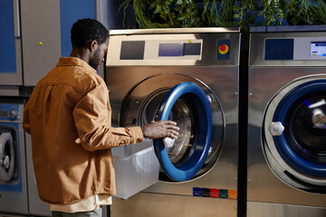 Young African American man opening round door of automatic washing machine before putting dirty...