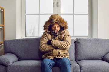 Freezing man in warm jacket sitting on sofa at home. Unhappy warmly dressed young man feeling cold...
