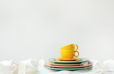 The image has a bright and minimalistic setting. A cup and saucer are placed on top of a stack of colorful plates - green and pink tones are visible.