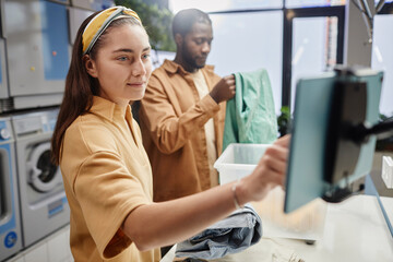 Obraz premium Young pretty client of laundry using tablet while weighing clothes after washing them in automatic machine and counting cost of service