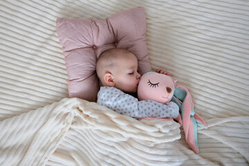 Baby with pink bunny soft toy. A girl of European appearance, wearing a light light jumpsuit, is sleeping in the crib. Sleep schedule, care and hygiene of a child up to one year old.