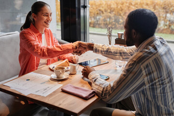 Happy young female manager shaking hand of African American male colleague over table with coffee,...
