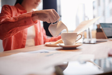 Young businesswoman in coral shirt pouring fresh milk or cream into cup of coffee while sitting by...