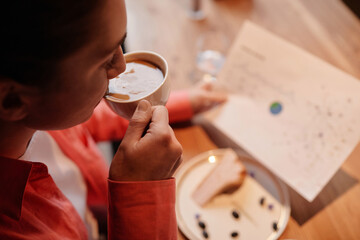 Close-up of young busy female economist having cup of cappuccino in cafe in the morning and looking through financial papers