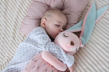 Baby with pink bunny soft toy. A girl of European appearance, wearing a light light jumpsuit, is sleeping in the crib. Sleep schedule, care and hygiene of a child up to one year old.